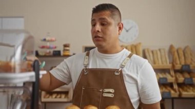 Genç İspanyol bir adam pastanede çalışıyor. Kapalı mekanlarda hamur işleri düzenliyor. Önlük takıyor ve arka planda ekmek somunları var.