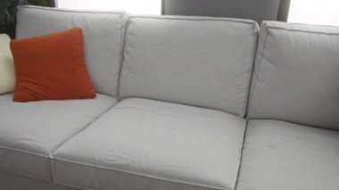 Rahat bir oturma odasında, gri bir koltukta oturan, renkli yastıklı, kestane rengi bir gömlek ve kot giyen, düşünceli ve sakin bir ev ortamında görünen bir adam..