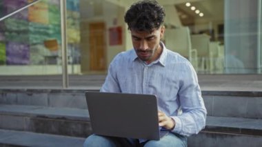 Şehir basamaklarında dizüstü bilgisayarlı genç bir adam mavi bir gömlek giyiyor ve kameraya gülümsüyor. Etrafı modern şehir mimarisi ve renkli duvar resimleriyle çevrili..