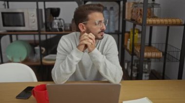 Ofis ortamında dizüstü bilgisayar kullanan gözlüklü genç adam, raflar ve mutfak eşyalarıyla çevrili, sıradan bir çalışma ortamı yaratıyor..