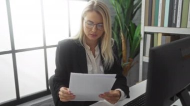 Modern bir ofiste doküman okuyan, sarı saçlı, gözlüklü, odaklanmış çalışma ortamını vurgulayan kadın doğal ışık ve profesyonel dekor kullanıyor..