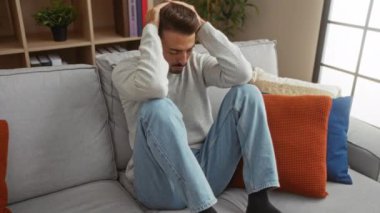 Genç adam rahat bir oturma odasında, renkli yastıklarla kanepede oturuyor. Stres ya da hayal kırıklığını ifade ediyor. Etrafı bitkilerle dolu kitaplıklarla çevrili..