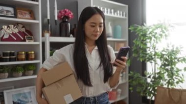 Genç Asyalı bir kadın karton bir kutu tutuyor ve güzel dekore edilmiş bir ev dekorasyonu dükkanında akıllı telefon kullanıyor ve arkasında çeşitli eşyalar sergileniyor.