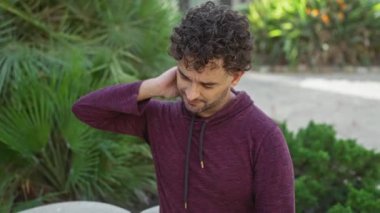 Bordo kapüşonlu genç adam yeşil şehir parkında rahatsız bir şekilde boynunu ovalıyor..