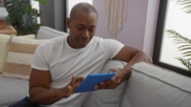 Genç adam rahat oturma odasındaki kanepede tablet kullanırken gülümsüyor, modern dekorla evde rahat bir atmosferin tadını çıkarıyor, kişisel alanların tadını çıkarıyor.
