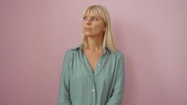 Orta yaşlı sarışın, tişörtlü, ciddi yüz ifadesi olan, doğal ve sade görünen, soyutlanmış pembe arka planı olan, kendine güvenen ve güvenen yaşlı bir kadın.