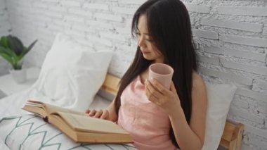 Bir fincan çayla yatakta kitap okuyan bir kadın, rahat bir yatak odası ortamında gevşeme sergiliyor tuğla duvar arkası ve doğal ışıklandırma ile