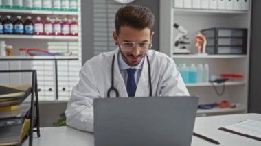 Genç İspanyol erkek doktor, bir hastane odasında tıbbi ekipman ve dosyalarla çevrili dizüstü bilgisayara bakarken yumruklarını sıkarak kutlama yapıyor..