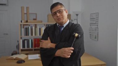 Elinde tokmak tutan İspanyol asıllı genç bir adam hukuk kitapları ve modern iç mimarisi olan bir ofiste güven içinde poz veriyor..