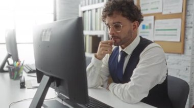 Ofisteki bir bilgisayarda çalışan genç adam, bir monitördeki verileri incelikle inceliyor. Etrafı dokümanlar ve ofis malzemeleriyle çevrili, profesyonel bir atmosferi aktarıyor..