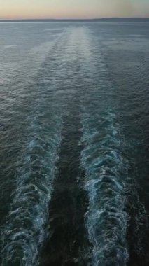 Okyanus dalgaları açık denizde gün batımında geminin arkasında süzülüyor. Renkli bir gökyüzünün altında uzak kıyıları olan sakin bir açık deniz manzarası yaratıyor..