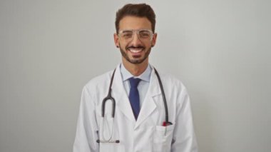 Beyaz önlüklü ve steteskoplu gülümseyen genç bir doktor izole edilmiş beyaz duvar arkaplanının üzerinde güven ve profesyonelliği işaret ediyor..