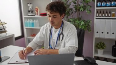 Klinikte steteskop takmış genç bir adam, tıbbi malzeme ve belgelerle çevrili bir dizüstü bilgisayar üzerinde çalışırken not alıyor..