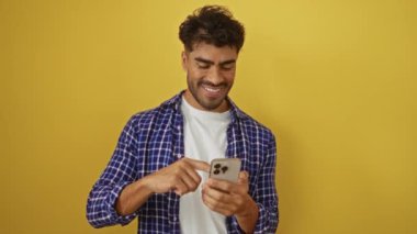 Genç adam canlı sarı arka plana karşı akıllı telefon kullanırken gülümsüyor, günlük ekose gömlek giyiyor ve modern saç stili ve sakallı İspanyol özellikleri sergiliyor.