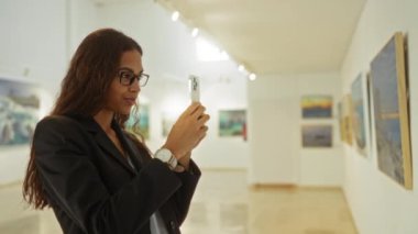 Kapalı bir galeri ortamında resim çeken bir kadın, modern sanatsal ifadelerin özünü ve sergi alanının atmosferini akıllı bir telefonla yakalıyor..