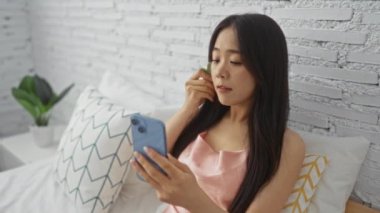 Yatak odasında dinlenen, mavi telefon kullanan, beyaz yastıklı ve yeşil bitkilerle cilt bakımı yapan bir kadın.