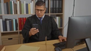 Ofisteki genç yargıç akıllı telefondan mesaj atıyor. Masasında bilgisayar ve kitaplıklarla oturuyor.