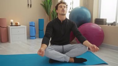 Spor salonundaki yoga minderinde meditasyon yapan bir adam egzersiz ekipmanları ve bitkilerle çevrili, dingin ve odaklanmış bir fitness ortamı yaratıyor..