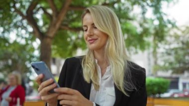 Şehir parkında akıllı telefon kullanan, iş elbiseli sarışın, şehir yeşilliğinin ortasında gülümseyen, gevşek ifadeyle online aktivitelere katılan bir kadın..