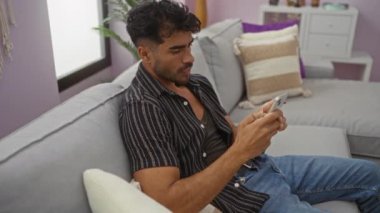 Genç adam, bir apartman dairesinde rahat bir kanepede rahat bir şekilde otururken akıllı telefona bakıyor, modern ve rahat bir yaşam tarzını somutlaştırıyor..