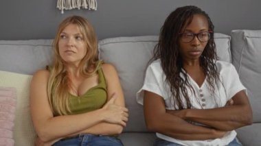 İki kadın evde kollarını kavuşturmuş, mutsuz ve mesafeli bir şekilde oturma odasında oturuyorlar.
