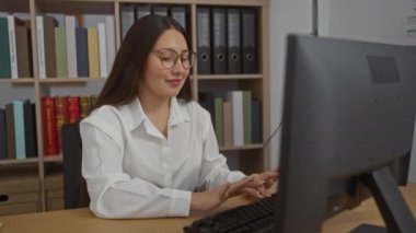 Ofiste çalışan bir kadın bilgisayarın arka planında kitap rafları olan.