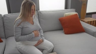 Hamile bir kadın rahat bir oturma odasında rahat bir koltukta oturuyor, nazikçe karnını tutuyor, modern bir apartman dairesinde sıcaklığını ve beklentilerini ifade ediyor..
