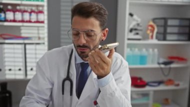 Tıbbi malzemeler ve anatomik modellerle çevrili iyi organize edilmiş bir klinikte steteskopla konuşan İspanyol erkek doktor dikkatle bir göreve odaklanıyor..