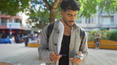 Şehir parkında duran sakallı genç adam telefonunu kontrol ediyor ve elinde şehir hayatı ile çevrili bir ağaç gölgesinde modern tarz ve güven yayıyor..