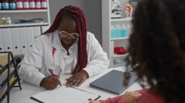 Gözlüklü doktor ve hasta, sağlık hizmetleri kliniğinde malzeme raflarıyla tıbbi notları tartışıyor..