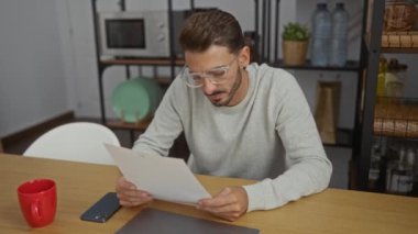 Ofiste masa başında evrak okuyan İspanyol bir adam, arka planda dizüstü bilgisayar, kahve ve mutfak aksesuarları olan, modern, rahat bir çalışma alanı sergiliyor..