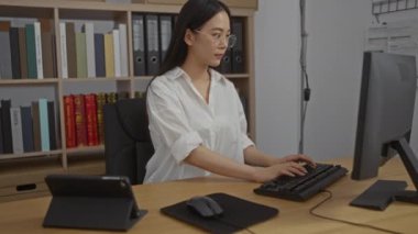 Çin 'de bir ofiste çalışan genç bir kadın bilgisayar ve tablet kullanarak arka planda kitaplarla dolu bir rafla.