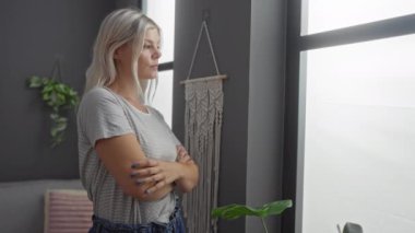Sarışın kadın oturma odasında kollarını kavuşturmuş pencereden dışarı bakıyor sakin bir günde makrome dekor ve yeşil bitkilerle içeri bakıyor.