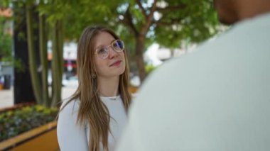 Kadın, şehir parkında, gün ışığında erkeklerle konuşuyor, gülümsüyor, ağaçlarla ve şehir arkaplanlarıyla meşgul oluyor, gündelik bir konuşma düzenliyor..