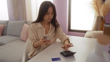 Oturma odasında telefon ve hesap makinesi kullanan genç Çinli bir kadın. Masadaki kredi kartıyla harcamaları hesaplıyor.