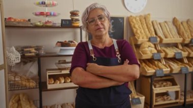 Bir pastanedeki İspanyol kadın, kollarını katlamış, taze ekmekle dolu rafların arasında duruyor, kapalı bir dükkânda olgun bir kadın güveni sergiliyor..