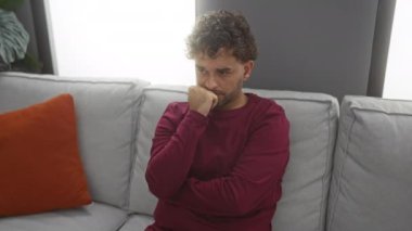 Genç adam modern bir oturma odasında portakal rengi yastıkla kanepede oturuyor eli çenesinde dalgın dalgın dalgın bakıyor..
