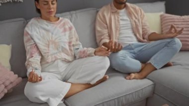 Çift, rahat bir oturma odasındaki kanepede birlikte meditasyon yapıyor. Uyumlu kıyafetlerle rahatlığı ve farkındalığı somutlaştırıyor.