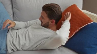 Genç bir adam evinde rahat bir oturma odasında rahat ve çekici bir yaşam tarzını sergileyerek bir kanepede dinleniyor..