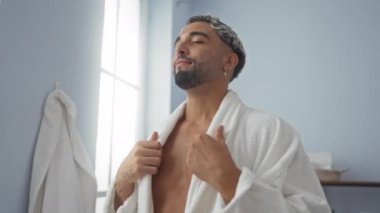 Genç, Arap, sakin bir kaplıcada, beyaz bir bornoz giyerken rahatlatıcı bir anın tadını çıkaran adam, doğal ışık altında, lüks bir sağlık merkezinde huzurlu bir ifadeyle.