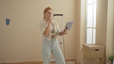 Ev yenilemeyi düşünen bir kadın dizüstü bilgisayar kullanıyor. Oturma odasında kutular ve merdivenlerle dikiliyor. Bir hareket öneriyor ya da karar değiştiriyor..