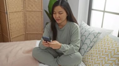 Hoş olmayan bir yüz ifadesiyle telefona bakan kadın yatak odasındaki yastıklarla yatağında oturuyor..