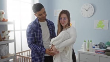 Bebek eşyaları ve yumuşak ışıklandırmayla çevrili sıcak bir yatak odasında bebeklerini kucaklayan çift sıcak bir aile ortamı yaratıyor..