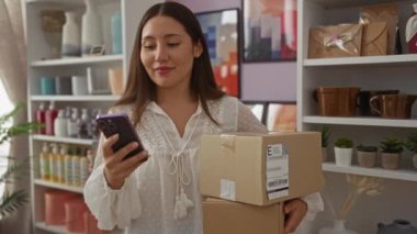 Dekoratif eşyalar ve bitkilerle dolu raflarla çevrili bir mağazada paketleri tutan ve akıllı telefon kullanan bir kadın.