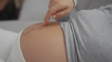 Hamile kadın rahat bir evde karnına hafifçe dokunuyor. Modern bir dairede annelik beklentisinin samimi ve huzurlu anlarını anlatıyor..
