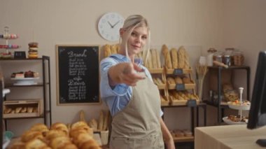 Kameraya el kol hareketi yapan genç bir kadın. Kapalı mekandaki fırında elleri ve arka planda ekmek ve hamur işleri olan bir kadın.