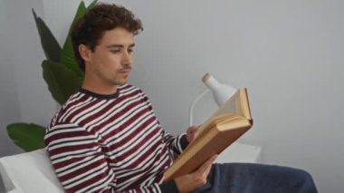 Çizgili süveter giyen genç adam rahat yatak odasında, beyaz lamba ve yeşil bitkiyle kitap okuyor. Huzurlu ve rahatlatıcı bir iç mekan yaratıyor.