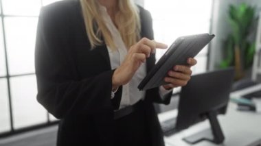 Modern ofiste gülümseyen tabletli bir kadın, gözlüklü ve siyah takım elbiseli, pencere ışığıyla aydınlatılmış sarı saçlı, profesyonel teknoloji kullanımı ve iş ortamını tasvir ediyor..