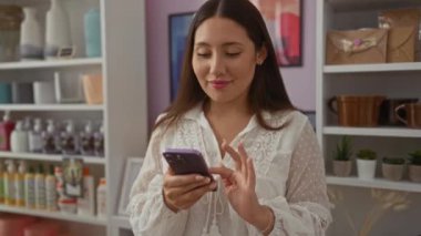 Arka planda çeşitli dekoratif eşyalar, vazolar ve güzellik ürünleriyle dolu rafları olan bir ev dekorasyonu mağazasında akıllı telefon kullanan genç bir kadın..