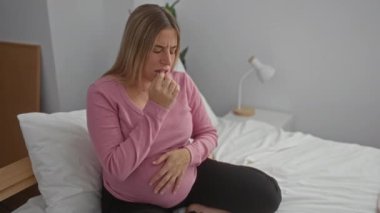 Hamile kadın rahat bir yatak odasında, eli karnının üstünde oturuyor. Rahatsızlığını dile getiriyor. Ev ortamında beklenti ve annelik hissi veriyor..
